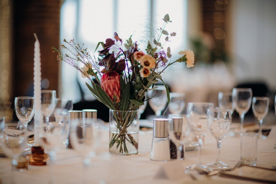 A floral arrangement featuring a variety of fresh flowers, including purple and cream blooms, arranged in a clear glass vase on a dining table. The bouquet contains a mix of flowers with different tex