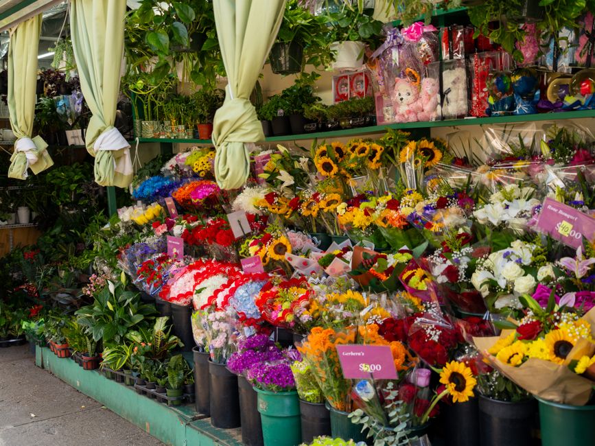 An outdoor display of various flower bouquets at a flower shop, with arrangements featuring sunflowers, roses, lilies, and other mixed flowers in vibrant reds, yellows, purples, and whites. The bouque