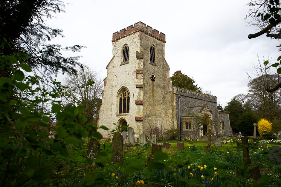 A traditional church with a tall, square tower featuring arched windows and a weathered stone façade, surrounded by a graveyard with weathered headstones and patches of green grass. In the foreground