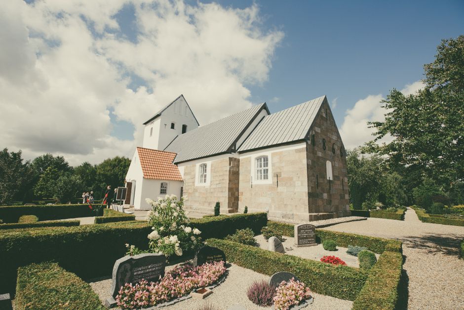 A serene outdoor scene showing a small historic stone church with a pointed roof and white walls, set against a partly cloudy sky. In the foreground, a well-maintained graveyard features several grave