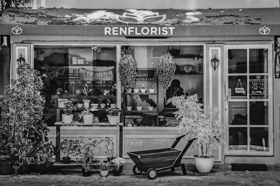 Black and white image of a flower shop named 'Renflorist' with large front windows displaying various potted plants and floral arrangements. Outside the shop, there are several potted plants and hangi