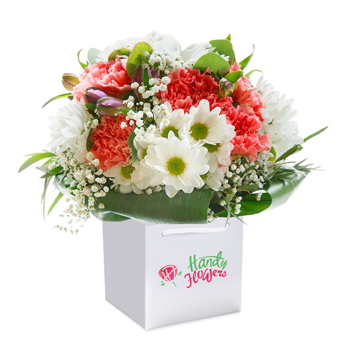 Presented upright in a sleek, square white gift bag, this vibrant floral arrangement beautifully embodies the spirit of Finsbury-a place where tradition and modernity dance together. The bouquet's composition is both abundant and refined: ruffled coral-pink and vivid red carnations set a lively pace, offset perfectly by crisp white daisy chrysanthemums whose lime-green centres seem to glimmer with cheerfulness. Dotted between these, airy tufts of white gypsophila soften the bouquet and provide a touch of springtime freshness. Tucked among the blooms on the left, you'll spot a few closed, purple-tinged buds, inviting a hint of anticipation. Lush, broad green leaves form a dramatic collar at the base, their glossy surfaces reflecting gentle light-a tribute to the leafy enclaves and hidden gardens found throughout Finsbury. The white bag, with sturdy ribbons for easy carrying, ensures the bouquet arrives as immaculate as when it left the local florist's workbench, ready to grace a doorstep on St. John Street or a gathering in Clerkenwell. Set against a pure white background, the arrangement radiates with vitality and artisanal attention to detail, highlighting the freshness and premium craftsmanship so characteristic of flower delivery in this part of London. Ideal for birthdays, anniversaries, or spontaneous expressions of care, this bouquet captures the artistic flair and personal touch of Finsbury's dedicated florists.